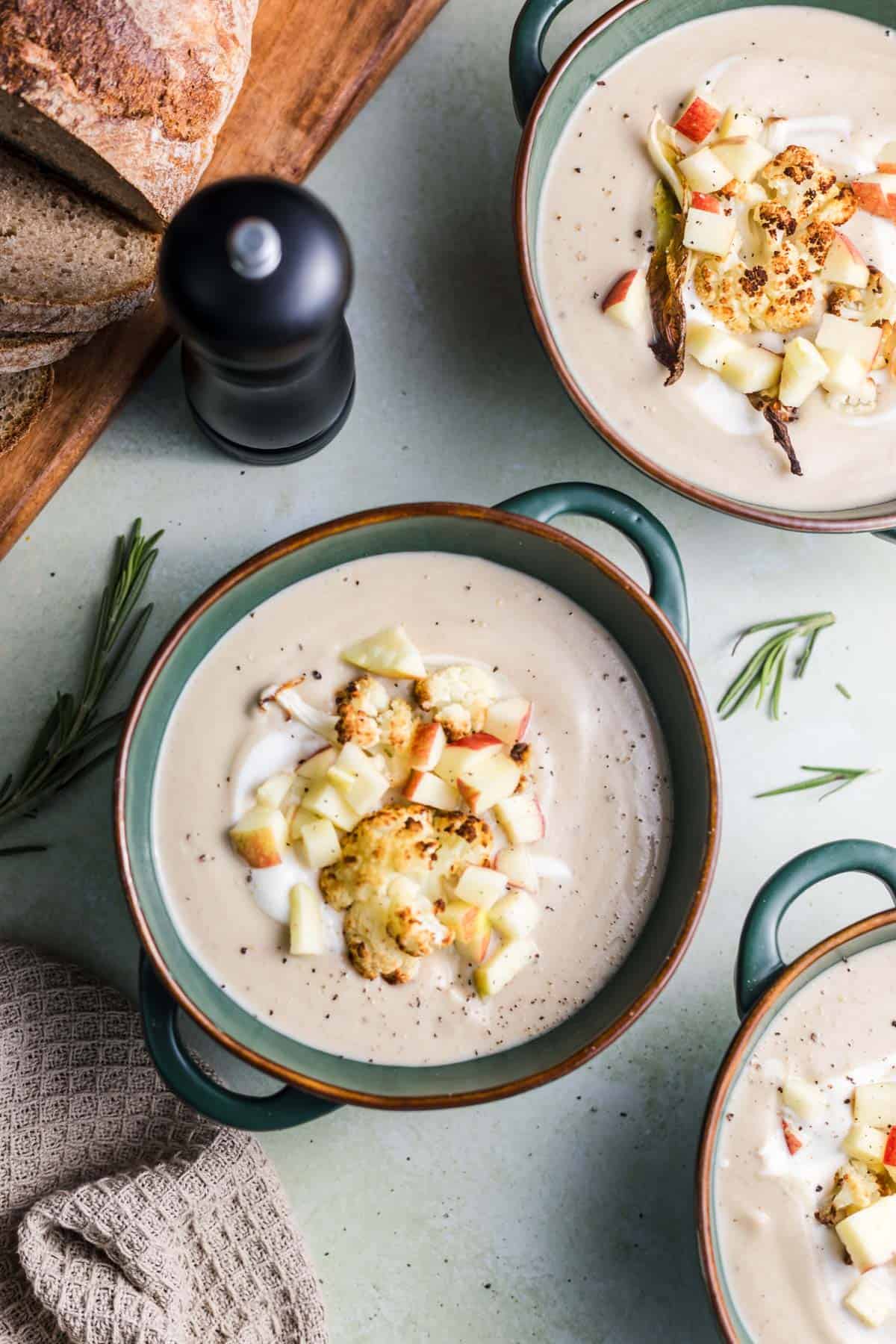 Bowls of creamy roasted cauliflower soup with chestnuts and cashews, served with bread and fresh rosemary