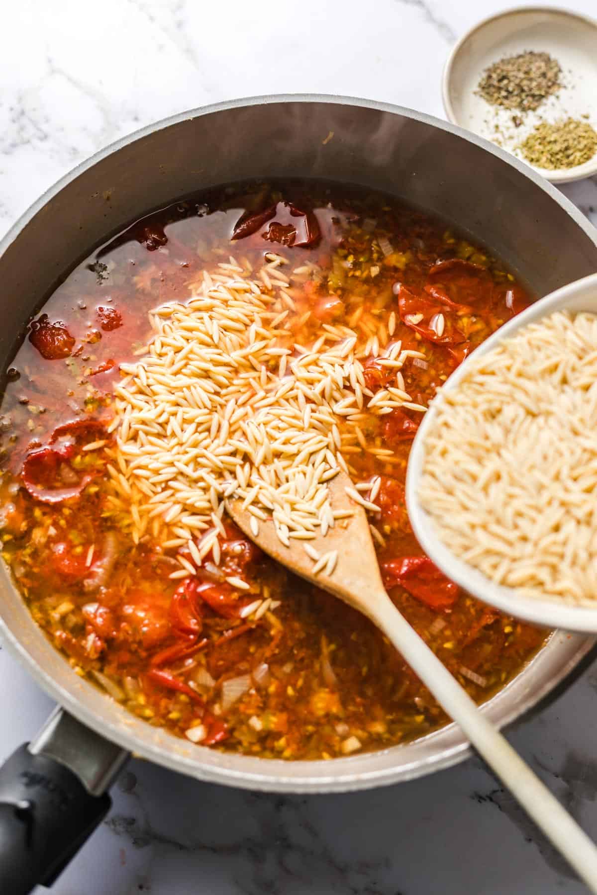 Stirring orzo pasta into the tomato broth with dried herbs