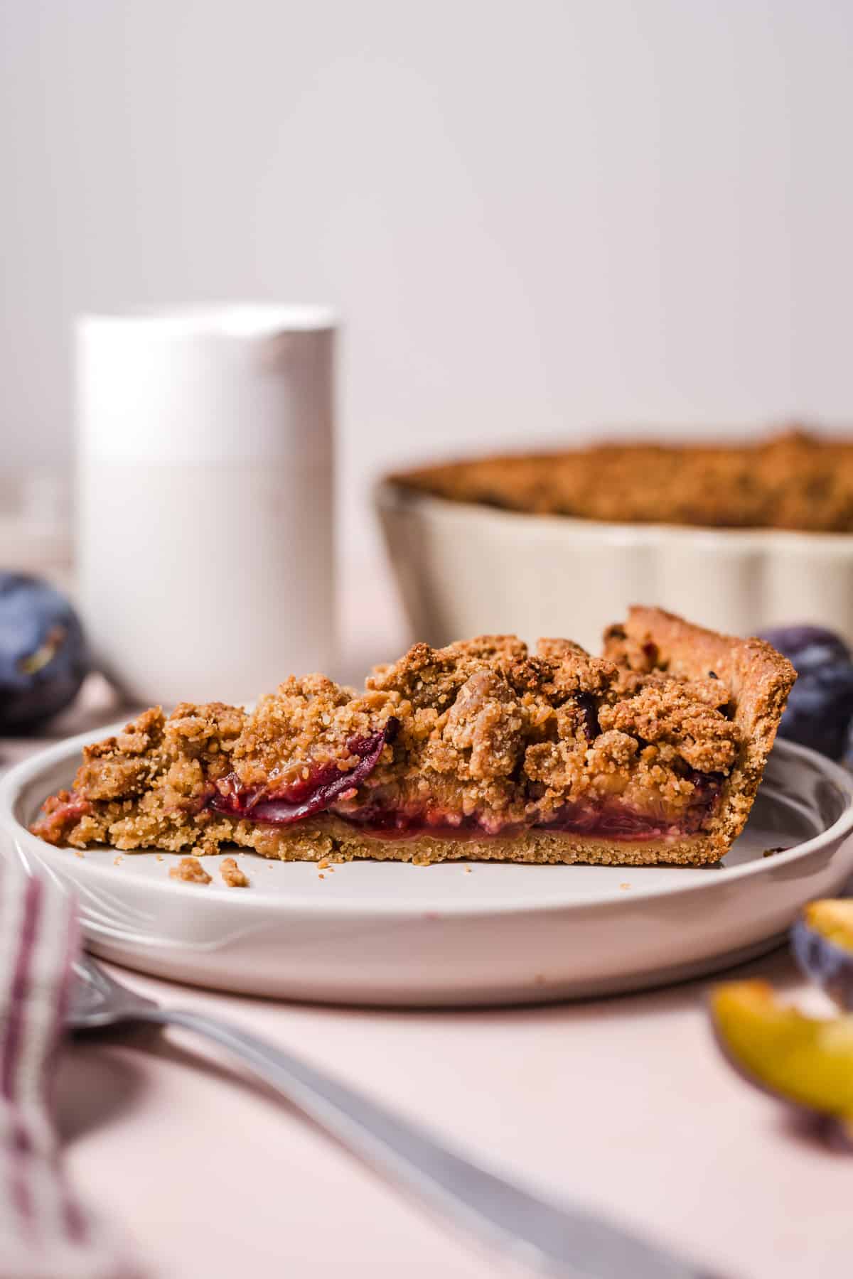 Slice of vegan plum crumble pie made with cashew butter and date sugar, whole pie in the background